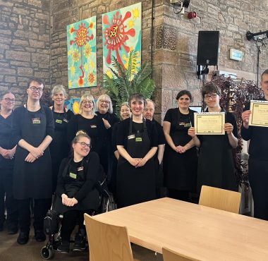 2 volunteers to the right of the photo wearing The Usual Place uniform and holding a certificate each. 14 people stand around them, 3 are volunteers and 11 are mentors and staff. They are all looking into the camera and smiling.