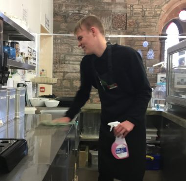 A volunteer standing centre of the photo wearing The Usual Place uniform. He is concentrated on wiping the counter with a cloth and a spray bottle, cleaning as he goes.