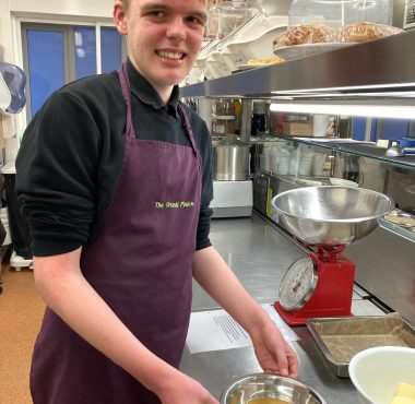 A volunteer standing to the left of the photo wearing The Usual Place kitchen uniform. He is holding a bowl of cracked eggs ready to bake. He is looking into the camera and smiling.