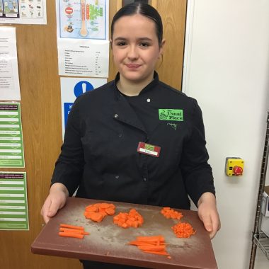 A volunteer with brown hair wearing The Usual Place kitchen uniform. She is in the kitchen, holding a cutting board with chopped carrots on it. She is looking into the camera and smiling.
