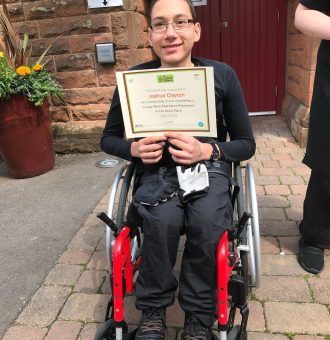 A volunteer in a wheelchair in the centre of the photo wearing The Usual Place uniform. The entrance to The Usual Place is behind him. He is holding a certificate, looking into the camera and smiling.