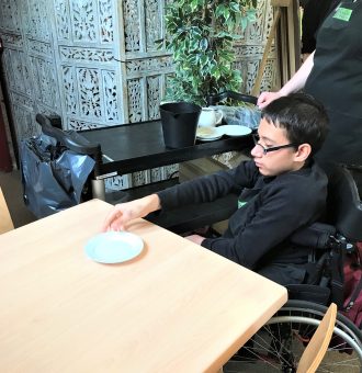 A volunteer in a wheelchair in the centre-right of the photo. He is focused on helping to clear the table, carrying a plate to the cleaning trolley beside him.