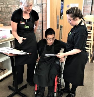 A volunteer in a wheelchair in the centre of the photo with another volunteer to his right and a mentor standing to his left. They are all wearing The Usual Place uniform. They are focused on a clipboard, ready to welcome guests and customers.