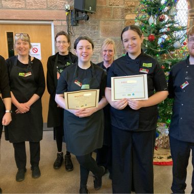 2 young volunteers centre of the photo wearing The Usual Place uniform. Behind them are 4 mentors and 1 volunteer, wearing The Usual Place uniform. The volunteers centre of the photo are holding a certificate each. All of them are smiling into the camera.