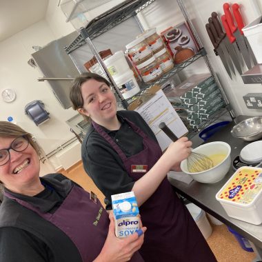 A volunteer centre of the photo and a mentor to the left of the photo wearing The Usual Place kitchen uniform. The volunteer is mixing cake batter together while the mentor holds flour ready for the next step in baking. They are both looking into the camera and smiling. The photo is tilted diagonally.