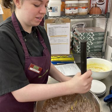 A volunteer with brown hair wearing The Usual Place kitchen uniform. She is concentrated on stirring chocolate cake mixture in a bowl.