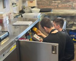 A volunteer centre-right of the photo wearing The Usual Place uniform. There is another volunteer next to him, also wearing The Usual Place uniform. They are turned away from the camera, checking stock under the counter.