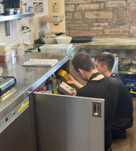 A volunteer centre-right of the photo wearing The Usual Place uniform. There is another volunteer next to him, also wearing The Usual Place uniform. They are turned away from the camera, checking stock under the counter.