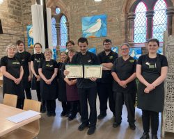 One of the volunteers standing in the centre of the photo wearing The Usual Place uniform. He is holding 2 certificates. Behind him are 3 other volunteers and 7 mentors and staff, also wearing The Usual Place uniform. They are all looking into the camera and smiling.