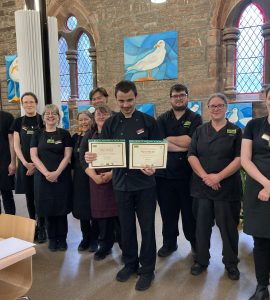 One of the volunteers standing in the centre of the photo wearing The Usual Place uniform. He is holding 2 certificates. Behind him are 3 other volunteers and 7 mentors and staff, also wearing The Usual Place uniform. They are all looking into the camera and smiling.