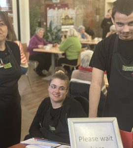 A volunteer to the right of the photo wearing The Usual Place uniform. To the left of him are another volunteer and a mentor, also wearing The Usual Place uniform. They are ready to welcome customers into the cafe and show them to their tables. They are all looking into the camera and smiling.