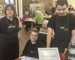 A volunteer to the right of the photo wearing The Usual Place uniform. To the left of him are another volunteer and a mentor, also wearing The Usual Place uniform. They are ready to welcome customers into the cafe and show them to their tables. They are all looking into the camera and smiling.