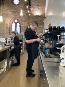 A volunteer in the centre of the image is using a Barista machine. He is holding a jug of milk under a pipe coming form the machine. A mentor is stood behind him to the left, checking a slip of paper.