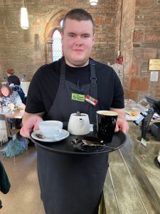 A volunteer is carrying a tray with a coffee mug, a small teapot, a jug of milk, and a teacup. The volunteer is looking at the camera and smiling.