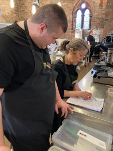 A mentor is looking down at a clipboard. She is holding a pen and is running through items on the clipboard. A volunteer stands in front of her. He is watching as the mentor goes through the checklist.