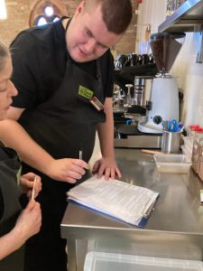 A mentor is looking down at a clipboard. She and a volunteer, stood behind her, are holding pens and are ticking off items on the clipboard. The volunteer is looking at the mentor and smiling at her.