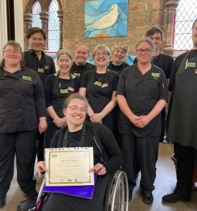 A volunteer in a wheelchair is sat centre of the photo wearing The Usual Place uniform holding a certificate for food safety and hygiene. Behind her stands 5 mentors and 3 volunteers, also wearing The Usual Place uniform. They are all looking into the camera and smiling.