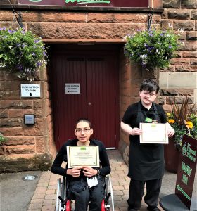 A volunteer with a wheelchair is sitting centre of the photo wearing The Usual Place uniform. Stood beside him is another volunteer. They are both holding food, health and hygiene certificates and smiling into the camera.
