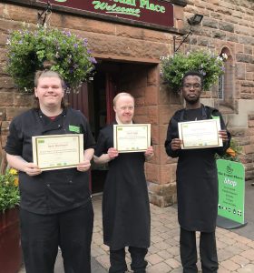 3 volunteers stood from the left to the centre and right of the photo wearing their Usual Place uniforms. They are all holding food, health and hygiene certificates and smiling into the camera.