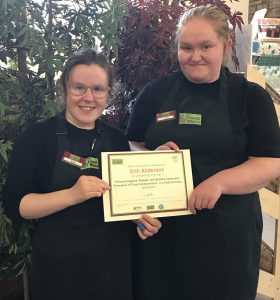 A volunteer with glasses standing to the right of the photo wearing The Usual Place uniform. A mentor is stood next to her, also wearing The Usual Place uniform. They are both holding a certificate that is being presented to the volunteer and smiling into the camera.