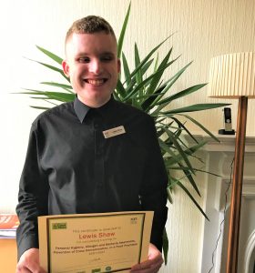 A volunteer stood to the centre-left wearing a black suit with a name tag. He is holding a certificate for food, health and hygiene and smiling.