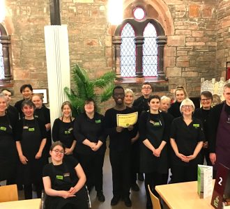 A volunteer stands in the centre of the photo wearing The Usual Place uniform. Around him are 6 volunteers and 9 mentors also dressed in The Usual Place uniform. They are all looking into the camera and smiling.