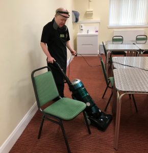 A volunteer standing centre-left of the photo wearing The Usual Place uniform. He is focused on hoovering the carpet in one of the meeting rooms.