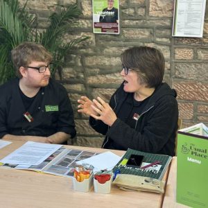 A volunteer sitting centre-left facing a mentor who is sitting centre of the photo. The volunteer is wearing The Usual Place kitchen uniform. The mentor is wearing a black jacket over her Usual Place uniform. On the table in front of them is paper work and documents relating to food health and safety.