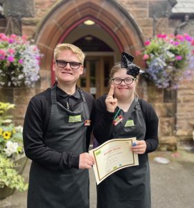 A volunteer with glasses and a bow stood centre-right in the picture wearing The Usual Place uniform. Stood next to her centre-left in the picture is another volunteer presenting a certificate for food, health and hygiene to her. They are both looking into the camera and smiling.