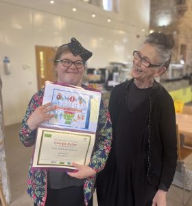 A volunteer with glasses and a bow stood centre-left in the picture holding a food health and hygiene certificate. Stood next to her centre-right in the picture is a mentor dressed in The Usual Place uniform. The volunteer is smiling at the camera and the mentor is smiling at the volunteer.
