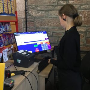 A volunteer with brown hair tied up in a bun wearing The Usual Place uniform and standing centre-right of the photo. She is looking away at the computer screen which is a till and finding out the total of the customer's food and drink.