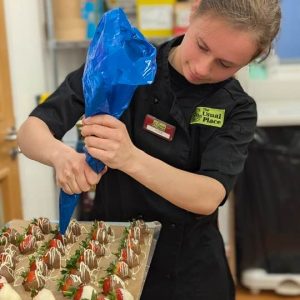 One of the volunteers with brown hair tied up in a bun wearing The Usual Place kitchen uniform. She is busy decorating canapés (milk and white chocolate covered strawberries), concentrating on piping the icing onto each strawberry.