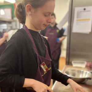 One of the volunteers with brown hair tied up in a bun wearing The Usual Place kitchen uniform. She is concentrated on holding a knife, about to cut a mushroom on a wooden chopping board to be used in a soup.