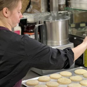 One of the volunteers with brown hair tied up in a bun wearing The Usual Place kitchen uniform. In front of her is a tray of shortbread biscuits and she is turned away from the camera, currently in the process of sprinkling a light dusting of sugar over them.