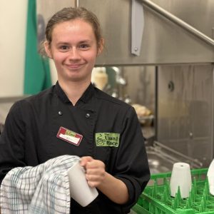 A volunteer with brown hair tied up in a bun wearing The Usual Place kitchen uniform. She is standing centre-left of the photo beside the washing up station, cleaning a mug and smiling into the camera.