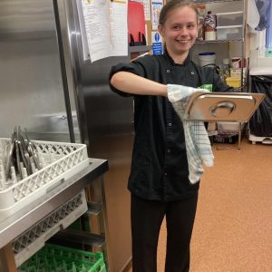 One of the volunteers with brown hair tied up wearing The Usual Place kitchen uniform. She is standing centre-right of the photo next to the washing up station, cleaning a metal tray and smiling into the camera.