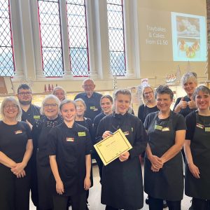 A volunteer with brown hair tied up in a bun standing centre-left of the photo. She is surrounded by staff and other volunteers. One of the other volunteers is holding a certificate for her. They are all looking into the camera and smiling.