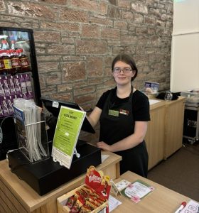 A volunteer with glasses standing centre in the picture wearing The Usual Place uniform. She is standing behind the welcome station using the register. She is looking into the camera and smiling.