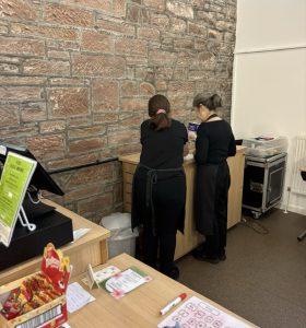 A volunteer with glasses standing centre of the picture wearing The Usual Place uniform. She is facing away from the camera looking over the booking sheets with a mentor, stood left of the picture, also wearing The Usual Place uniform.