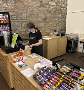 A volunteer with glasses standing centre-right of the picture wearing The Usual Place uniform. She is stood behind the welcome station using the register.