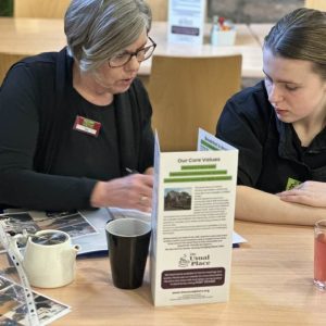A volunteer with brown hair is sat to the right of the photo at a table wearing The Usual Place uniform. She is filling out paper work and documents relating to food health and safety. Sat next to her centre-left of the photo is a mentor who is helping her make sense of the work. The mentor is talking to the volunteer here who is listening.