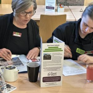 A volunteer with brown hair is sat to the right of the photo at a table wearing The Usual Place uniform. She is filling out paper work and documents relating to food health and safety. Sat next to her centre-left of the photo is a mentor who is helping her make sense of the work.