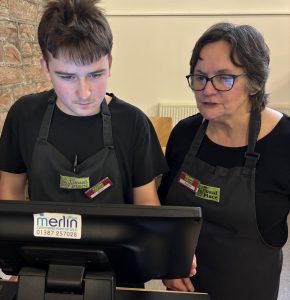 A volunteer with brown hair standing to the left of the photo wearing The Usual Place uniform. Standing next to him is a mentor with glasses who is also wearing The Usual Place uniform. They are looking at the welcome station's till screen, concentrated on putting the customer's order into the system.