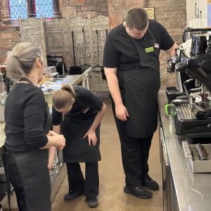 A volunteer with blonde hair to the left of the photo crouching down to put some dishes back from being cleaned. There is another volunteer to the centre-right of the photo operating the drinks machine. Standing to the left of the photo is a mentor. They are all wearing The Usual Place uniform and concentrated on what they are doing.