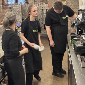 A volunteer with blonde hair to the centre-left of the photo putting some clean dishes away. There is another volunteer in the centre-right of the photo operating the drinks machine. A mentor stands to the left of the photo facing away from the camera. They are all wearing The Usual Place uniform and concentrated on what they are doing.