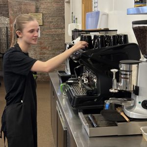 A volunteer with blonde hair tied back standing to the left of the photo wearing The Usual Place uniform and operating the drinks machine. She is not looking into the camera, instead concentrated on what she is doing.