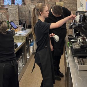 A volunteer with blonde hair standing centre of the photo and another volunteer with brown hair stood behind her. They are working at the Drinks and Cakes station, preparing orders for the customers. To the right of the photo is a mentor. They are all wearing The Usual Place uniforms.