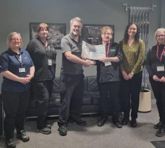 A volunteer stands with team members from Gates Dumfries and a mentor from The Usual Place. To the left of the photo are three of the team members, one of which the volunteer is holding a certificate of donation, and to the right of the photo is another team member and The Usual place mentor. The volunteer and mentor are wearing The Usual Place uniform and building passes around their necks. Everyone is looking into the camera and smiling.