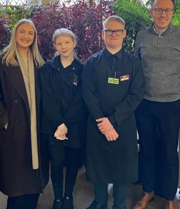 Two volunteers stand centre of the photo wearing The Usual Place uniform. On both sides of them are two team members from Farries Kirk & McVean who are wearing casual professional clothes. They are all looking into the camera and smiling.