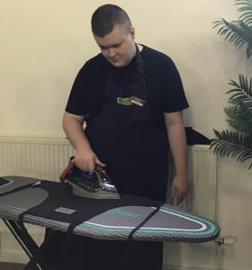A volunteer with black hair stood centre of the photo wearing The Usual Place uniform. He is ironing an apron on an ironing board. He is looking down and focused on what he is doing.